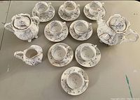 View of the porcelain tea set comprising a teapot, sugar bowl with lid, creamer, 8 teacups with matching saucers, arranged on neutral surface.