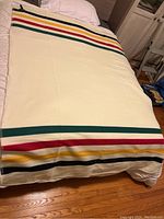 Cream colored fleece throw blanket with Hudson’s Bay multicolor stripes laid out on a bed.