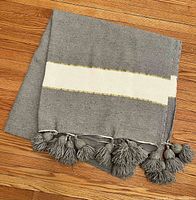 Folded view of the throw blanket showing grey color with a white stripe bordered by gold thread and tassels on the edge