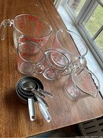 All Pyrex glass measuring cups and stainless steel measuring cups displayed on wooden tabletop showing sizes and handle details.
