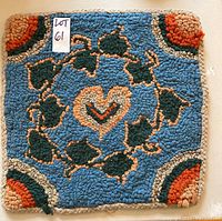 Top view of colorful hooked chair mat showing heart motif and leaf wreath on blue background with rounded corner patterns.