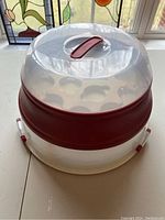 Front angle of Starfrit two-level cupcake and cake carrier showing clear lid with red handle locked onto base tier