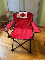 Red Canada Day branded folding camping chair with Canadian flag design on backrest, fabric seat and black steel frame, seen unfolded indoors.