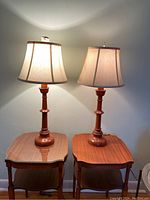 Pair of hand-turned pine candlestick lamps with fabric lampshades on wooden side tables, turned on and casting light on the wall.