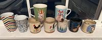 Front view of nine assorted mugs arranged in a row on a white windowsill showing various designs including animal faces, roosters, and colorful patterns.