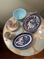 Top view of the blue and white Willow pattern serving dish, shallow Willow dish, small blue bowl and floral teacup on a glass table.