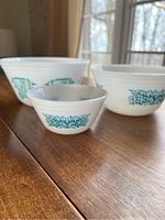 Three vintage opaque white glass mixing bowls sized approximately 11", 8", and 5" on a wooden surface, showing exterior blue patterned designs on each bowl.