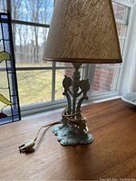 Small vintage painted metal lamp with decorative base and bird motifs under a beige textured lampshade, sitting on a wooden surface by a window.
