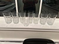 Seven clear water glasses of various sizes and designs arranged in a row on a window ledge, showing overall condition and size differences.