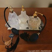 Front view of three cut glass perfume atomizer bottles displayed on a mirrored tray, two with black fabric bulbs and gold spray tops, one frosted glass with gold spray top.