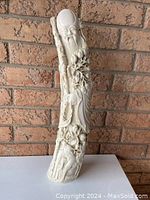 Front view showing detailed carved Asian figure holding staff with floral and animal motifs on elongated curved tusk-shaped resin sculpture.