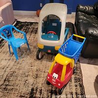 Blue plastic kids patio chair, Little Tykes Cozy Coupe ride-on car, and red/yellow shopping cart ride-on placed on carpet in living room.
