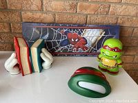 Photo showing Mickey Mouse hand bookends holding three books and next to them is a green Teenage Mutant Ninja Turtles Raphael statue and a green TMNT lamp base shaped like a mask. Behind these is a rectangular Spiderman wall print featuring Spiderman crawling among webs in blue tones.