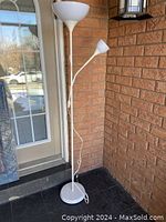 Full view of IKEA double floor lamp shown installed on porch against brick wall, displaying white circular base, main floor lamp, and smaller adjustable lamp branch with white shades.