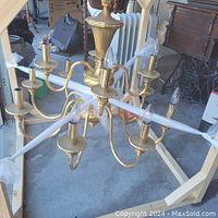 Full view of the brass chandelier hanging within a wooden frame on concrete floor. Shows eight arms curved outward with candle bulb holders and bulbs.