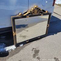 Full view of the black and gold ornate mirror leaning against a white vehicle outside, showing decorative top crest and corner accents.