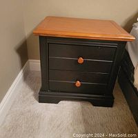 View of one nightstand showing dark grey painted body, two drawers with round orange-toned wooden knobs and wooden top with clear finish.