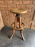 Full view of vintage wooden swivel stool showing round seat, four legs, and circular footrest against brick wall background.