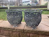 Pair of black decorative plant holders with ornate openwork design and curled feet placed outdoors on a brick ledge.