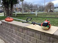 Side view of the full Husqvarna trimmer showing the orange trimmer head, metal shaft, and engine placement on a cardboard backdrop.