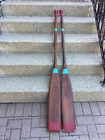 Two Mennonite wooden boat oars shown full length, propped on concrete steps. Red handles and tips, blue band near blade present, wood grain visible.
