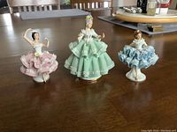 Three Dresden-style porcelain figurines on a wooden table: large green dress figurine in center, flanked by smaller pink and blue dress figurines