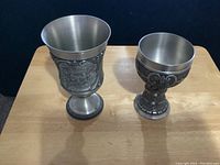 Two pewter goblets, one taller with heraldic design and one shorter with scrollwork design, placed on a wooden surface.