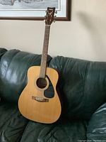 Front view of the acoustic guitar resting on a dark green leather couch showing the natural wood body, black pickguard, and neck with visible crack.