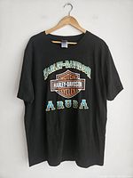 Front view of black Harley-Davidson T-shirt with Aruba print hanging on wooden hanger against white wall.