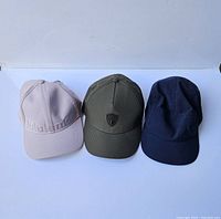 Front view of three caps in a row, light beige, olive green, and navy blue showing brand details and colors.