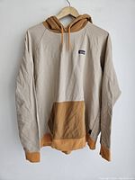 Full view of Patagonia hoodie hanging on wooden hanger, showing beige body with brown hood, front pocket, hem, and cuffs.