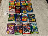 Photo showing all 16 sealed sports trading card packs laid out on white fabric background, including baseball, football, and hockey cards.
