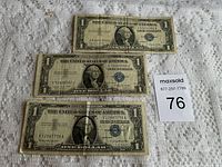 Three $1 US silver certificate banknotes arranged flat on a cloth surface with a lot number card visible.