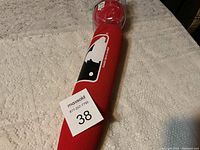Red foam toy baseball bat with Major League Baseball logo, lying on a beige textured fabric surface. Bat includes attached red toy ball wrapped in plastic.
