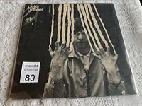 Front vinyl album cover showing Peter Gabriel with raised hands and striped effect, in protective plastic sleeve on tablecloth surface.