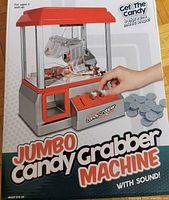 Front view of box showing toy claw machine in silver and red with candy inside and hand using controls. Includes gray plastic coins and emphasizes sound feature.