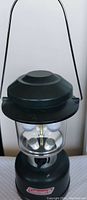 Front view of black Coleman outdoor lantern with carrying handle and batteries inside, showing the lantern switched off.