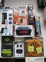 Photo of all seven items in original packaging arranged on a surface: Ice Cube Wizard ice cube maker, Cell Phone Umbrella holder, universal remote control, dual alarm clock radio, wireless ear pods, wireless charging pad, and mini dart board.