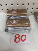 Two gold-colored Coors Light MC2015A power banks with included white USB charging cables, sealed in plastic wrap, placed on a shelf with a price label '80' visible.