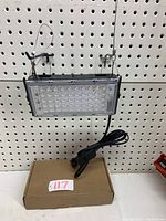 Front view of rectangular LED grow light hanging on wire cables with power cord coiled below and brown cardboard box beneath.