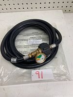 Full view of coiled black 12-foot propane hose with attached brass pressure gauge and fittings, laid on a pegboard and plastic.