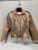 Front view of tan leather fringe coat with collar and fringe detailing on chest and arms, hung on pegboard.