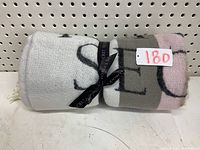 Rolled Victoria's Secret throw blanket tied with a black branded ribbon, showing color sections of white, gray, and pink with large black printed letters.