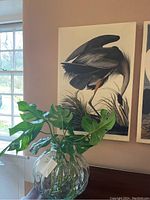 Canvas art depicting a Great Blue Heron next to a window with a glass vase of monstera leaves on a table beneath it.