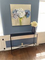 Black metal console table against blue wall beneath floral artwork, black hammered vase with faux white hydrangeas on top.