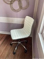 Photo of a white desk chair with black piping and chrome wheeled base, placed against a lavender wall near a window.