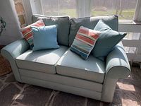 Front view of pastel green loveseat with four pillows, positioned in sunroom with stone flooring and large windows.