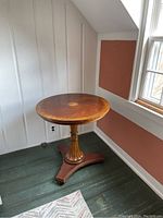 Front angle image of round wooden pedestal table showing carved pedestal base and tripoid platform base with decorative details, located in corner room.
