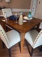 Photo of the Ethan Allen solid wood square table with medium brown finish. Table surrounded by upholstered cream curtains chairs with nail head trim. The table surface holds glassware and plates.