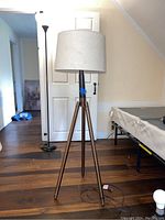 Full view of the bronze tripod floor lamp with white shade placed on wooden floor in a bedroom.